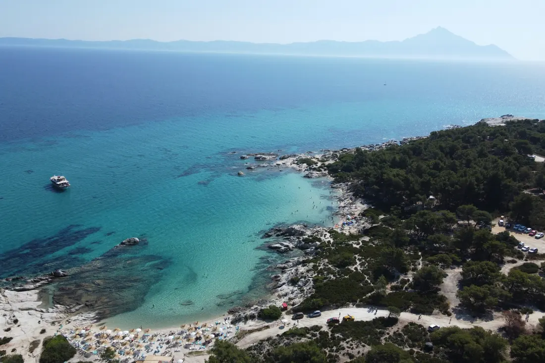 Aerial view of Kavourotrypes: pale rocks, small coves, parasols packed side by side and a hillside car park, with Mount Athos on the horizon